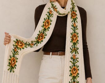 May include: Cream-colored scarf with a floral pattern featuring orange and yellow flowers with green leaves. The scarf has a fringed edge and is displayed against a neutral background.