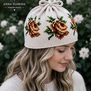 May include: Cream-colored knit hat with a drawstring closure and floral embroidery. The hat features brown, yellow, and green roses. The text "ANNA FLORIDA - TIMELESS BEAUTY" is printed on the hat. The hat is a winter accessory.