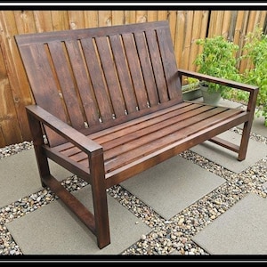 Può includere: Un divano da esterno in legno marrone scuro con schienale e seduta a doghe. Il divano è posizionato su un patio con piastrelle quadrate. Il testo "HOME MADE OUTDOOR SOFA PLANS" è visualizzato.