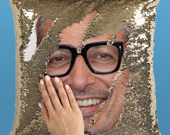Puede incluir: Un cojín dorado con lentejuelas con una imagen oculta revelada. La imagen muestra a una persona con gafas negras. Una mano está tocando la imagen revelada. El cojín está sobre un fondo azul.