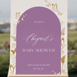 Puede incluir: Un cartel de bienvenida morado y blanco con mariposas doradas y el texto "Bienvenido al baby shower de Abigail. Estamos encantados de que est&eacute;s aqu&iacute;. 10.15.23".