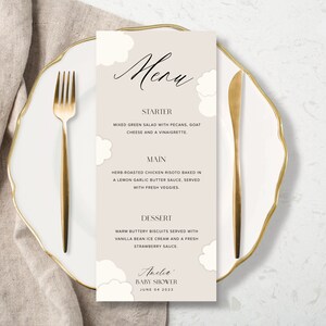 May include: A beige menu card with a cloud design. The menu lists starter, main, and dessert options for a baby shower. The text at the bottom reads "Amelia Baby Shower June 04 2023".