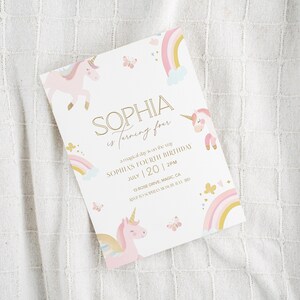 May include: A white invitation card with a unicorn theme. The card features a pink unicorn, rainbows, and butterflies. The text reads "SOPHIA is turning four" with the date July 20 and the time 2 PM.
