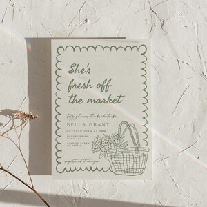 May include: A light-colored invitation with a scalloped border and the text "She's fresh off the market." Below, it reads "let's shower the bride-to-be BELLA GRANT." A drawing of a basket with flowers is included.