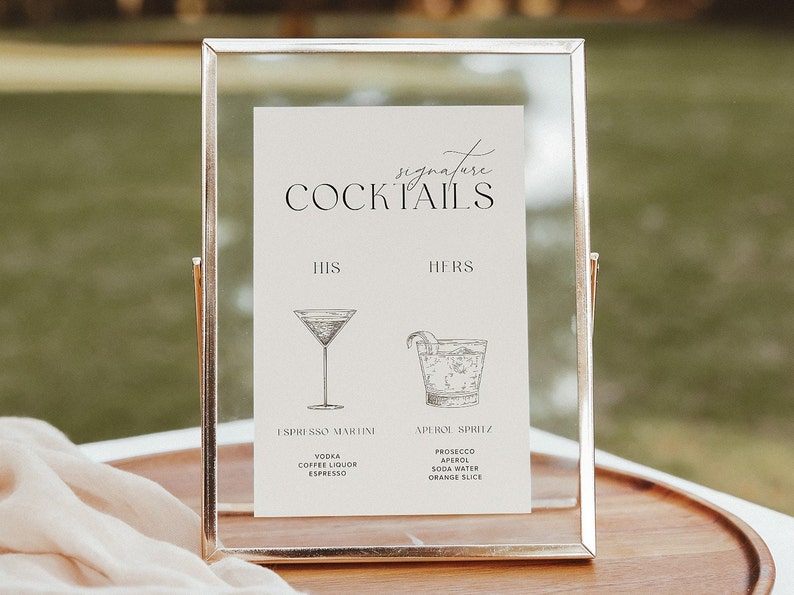 Peut inclure: Une pancarte dor&eacute;e avec le texte "signature COCKTAILS" et deux recettes de cocktails, l'une intitul&eacute;e "HIS" et l'autre "HERS".