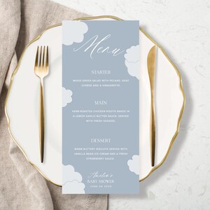 K&ouml;nnte beinhalten: Ein blaues und wei&szlig;es Baby Shower Men&uuml; mit einem Wolken-Design. Das Men&uuml; listet Vorspeise, Hauptgericht und Dessert auf. Der Text unten lautet "Amelie's Baby Shower June 04 2023".
