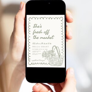 May include: A smartphone displays a bridal shower invitation with the text "She's fresh off the market." The invitation features a basket with flowers and text details in a decorative frame. The phone is held by two hands.