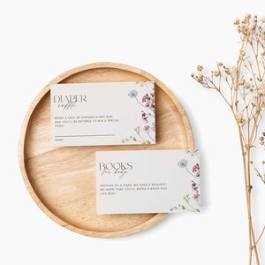 May include: Two white cards with floral designs and text. The top card says "Diaper Raffle" and asks guests to bring a pack of nappies for a chance to win a prize. The bottom card says "Books for Baby" and asks guests to bring a book instead of a card.