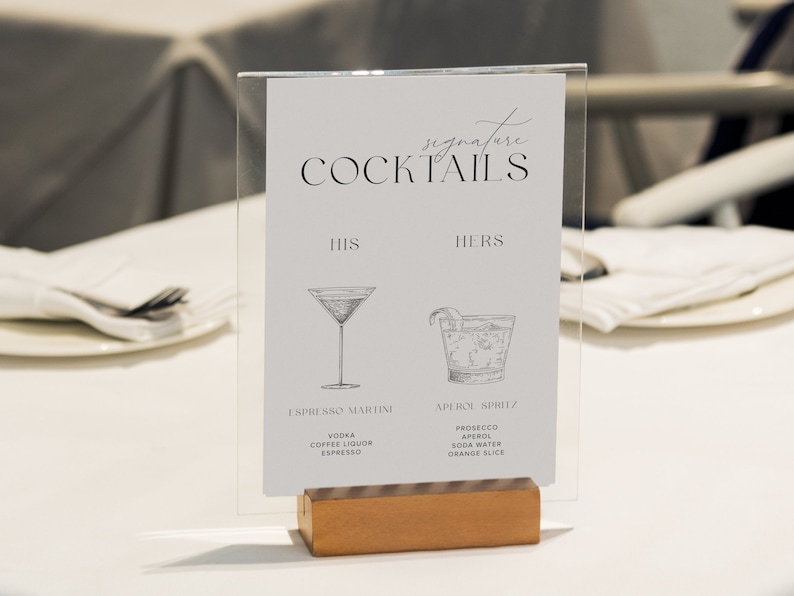 Peut inclure: Une pancarte en acrylique transparente avec une base en bois, affichant un menu de cocktails de mariage. La pancarte indique "Signature Cocktails" avec les titres "Son" et "Sa". Le cocktail "Son" est un Espresso Martini avec les ingr&eacute;dients suivants : Vodka, Liqueur de caf&eacute;, Espresso. Le cocktail "Sa" est un Aperol Spritz avec les ingr&eacute;dients suivants : Prosecco, Aperol, Soda Water, Tranche d'orange.
