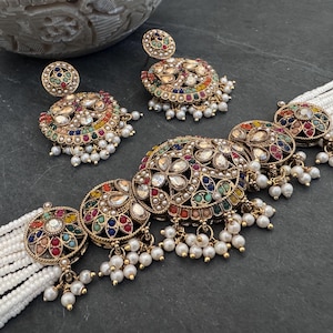 May include: A gold-toned necklace and earring set featuring circular motifs adorned with multi-colored stones and clear crystals. The necklace includes multiple strands of small white beads and dangling white pearl-like accents on each circular piece.