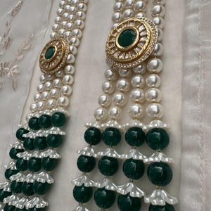 May include: Elegant pearl necklace with three strands, featuring gold-toned circular accents with green center stones and clear rhinestones. The necklace has alternating sections of large, dark green beads and white, flower-shaped accents.