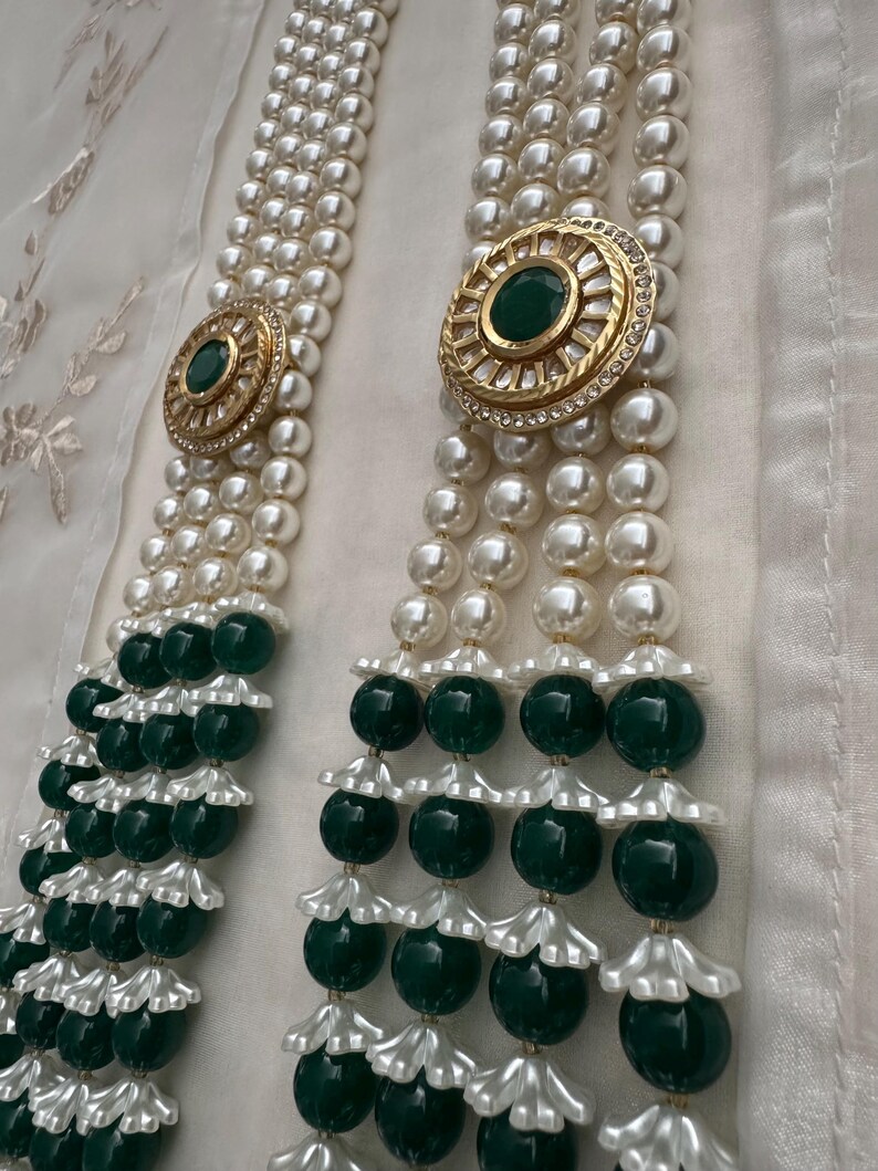 May include: Elegant pearl necklace with three strands, featuring gold-toned circular accents with green center stones and clear rhinestones. The necklace has alternating sections of large, dark green beads and white, flower-shaped accents.