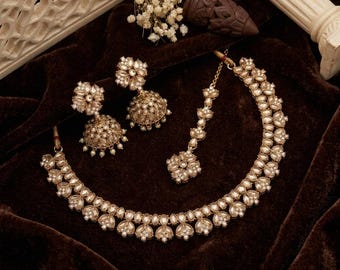 May include: A gold-toned jewelry set featuring a necklace with two rows of teardrop-shaped stones, a pair of bell-shaped jhumka earrings with intricate metalwork and small white beads, and a matching maang tikka with a floral-shaped pendant.