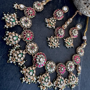 May include: A gold-toned necklace and earring set featuring floral-shaped motifs with inlaid red and green enamel, clear stones, and small white bead dangles. The jewelry pieces are connected by a gold cord with white bead accents.