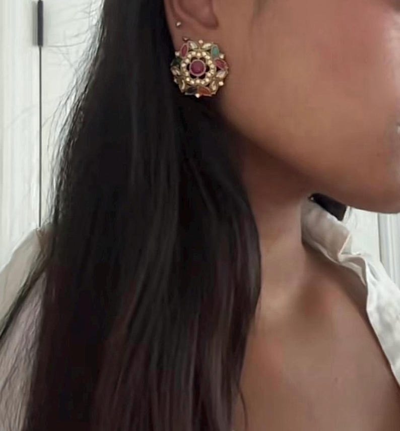 May include: A gold-toned, floral-shaped earring with a central ruby-colored stone, surrounded by smaller green, red, and white stones. The earring has a pearl-like border. The earring is worn on the ear.
