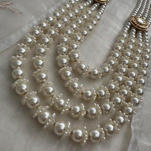 May include: A multi-strand pearl necklace with a gold-toned clasp. The necklace features multiple strands of white pearls, with decorative floral accents between the larger pearls. The clasp has a circular design with a central stone.