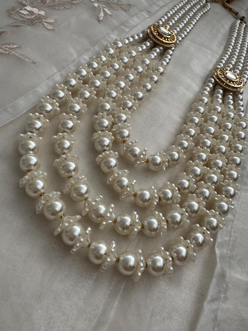 May include: A multi-strand pearl necklace with a gold-toned clasp. The necklace features multiple strands of white pearls, with decorative floral accents between the larger pearls. The clasp has a circular design with a central stone.