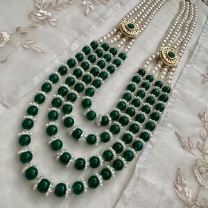 May include: A multi-strand necklace featuring alternating strands of green beads and white pearls. The necklace has gold-colored circular clasps with a green stone in the center. The necklace is displayed on a white, textured surface.