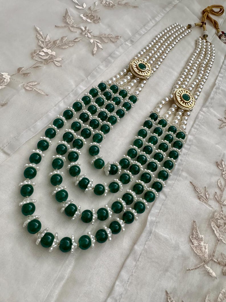 May include: A multi-strand necklace featuring alternating strands of green beads and white pearls. The necklace has gold-colored circular clasps with a green stone in the center. The necklace is displayed on a white, textured surface.