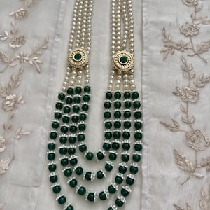 May include: A multi-strand necklace featuring white pearls and dark green beads. The necklace has gold-colored circular accents with a green center. The strands are layered, creating a cascading effect, and are displayed on a white fabric.