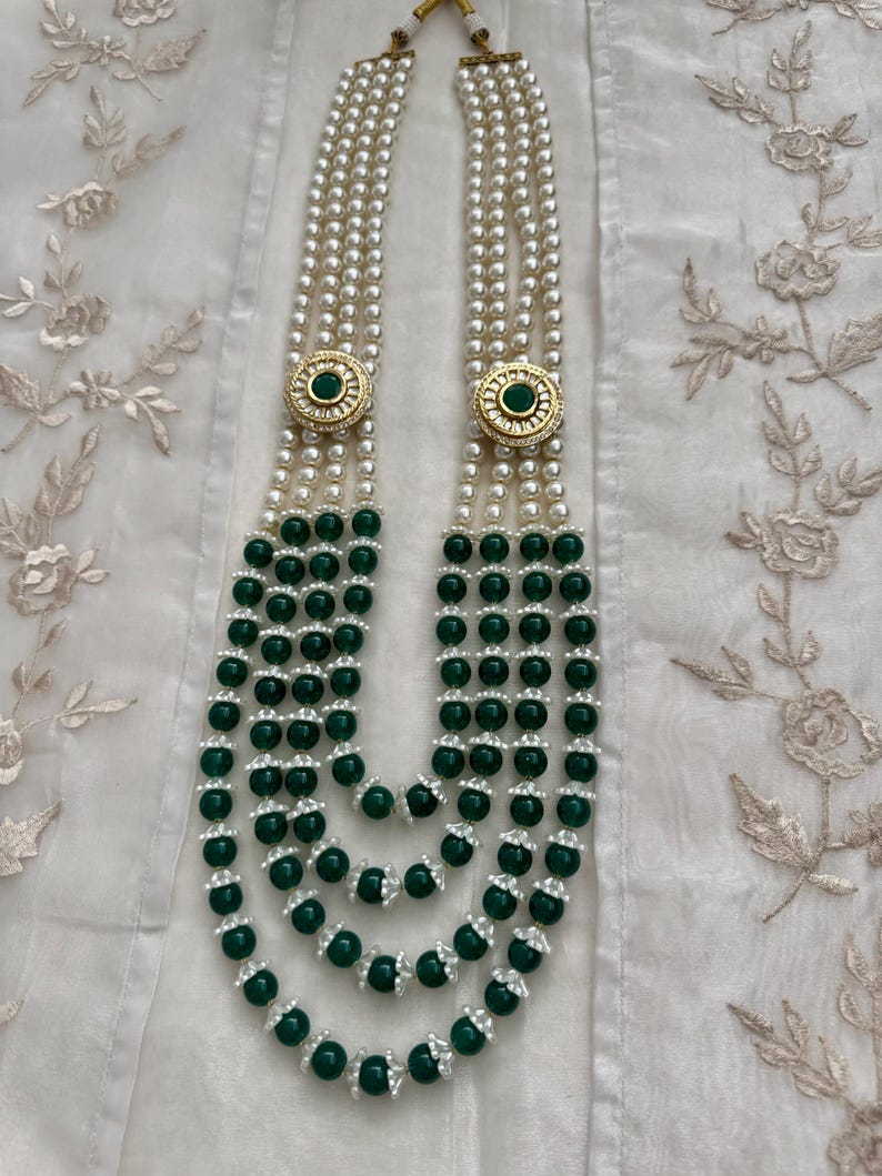 May include: A multi-strand necklace featuring white pearls and dark green beads. The necklace has gold-colored circular accents with a green center. The strands are layered, creating a cascading effect, and are displayed on a white fabric.