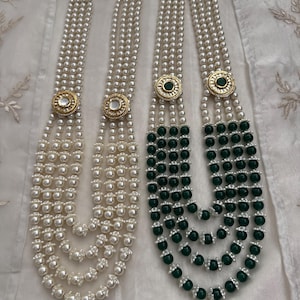 May include: Two necklaces with multiple strands of faux pearls. One necklace is all white pearls, and the other has green beads on the lower strands. Both necklaces have a gold-colored circular pendant with a central stone.