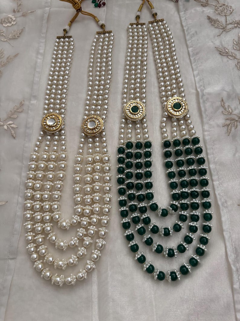 May include: Two necklaces with multiple strands of faux pearls. One necklace is all white pearls, and the other has green beads on the lower strands. Both necklaces have a gold-colored circular pendant with a central stone.