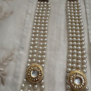 May include: A multi-strand pearl necklace with gold-toned circular pendants. The pendants are adorned with clear stones and a central clear gem. The necklace is displayed on a white fabric background.