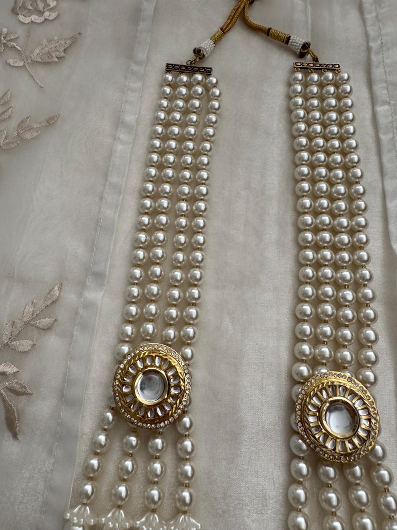 May include: A multi-strand pearl necklace with gold-toned circular pendants. The pendants are adorned with clear stones and a central clear gem. The necklace is displayed on a white fabric background.