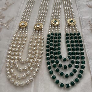 May include: Two necklaces with multiple strands of pearls. One necklace is all white pearls, and the other has green beads. Both necklaces have gold-colored circular clasps with decorative accents.