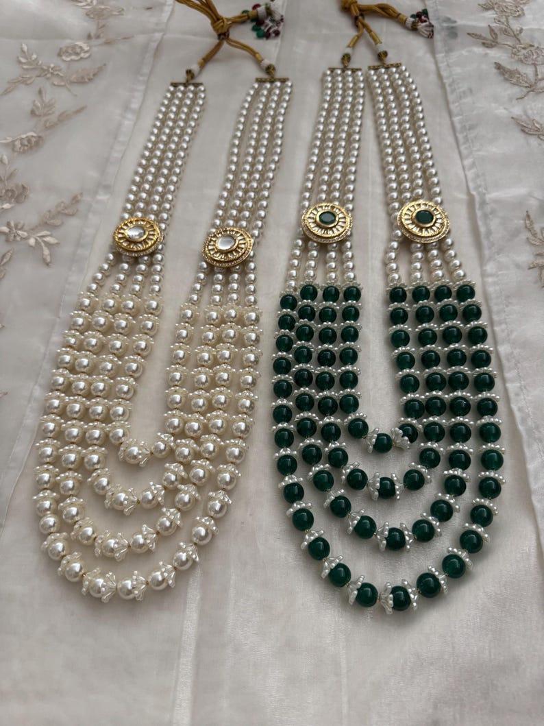 May include: Two necklaces with multiple strands of pearls. One necklace is all white pearls, and the other has green beads. Both necklaces have gold-colored circular clasps with decorative accents.