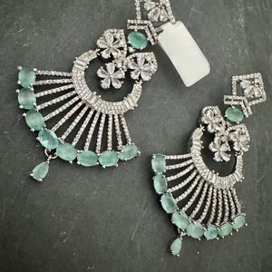 May include: A pair of silver-toned earrings with a fan-like design. Each earring features a square top with floral accents, a half-circle with clear stones, and a row of oval, light green stones. The earrings have a drop-style design.