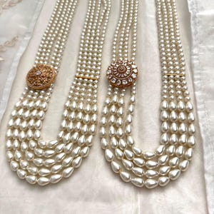 Peut inclure: Deux colliers de perles avec plusieurs rangs. Chaque collier présente un grand pendentif rond doré avec une pierre précieuse centrale et des pierres plus petites. Les rangs passent de petites perles rondes à de plus grandes perles en forme de goutte.