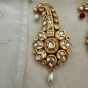 Puede incluir: Primer plano de un pendiente dorado y ornamentado con piedras claras de corte cuadrado y un colgante de perla en forma de lágrima. El pendiente presenta un diseño floral con una parte superior curvada, sobre un fondo blanco.