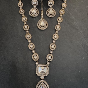 May include: A gold-toned jewelry set consisting of a long necklace, a pair of earrings, and a maang tikka. Each piece features clear, faceted stones set in ornate metal frames, with the necklace showcasing a large rectangular and teardrop pendant.