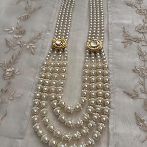 May include: A multi-strand pearl necklace with gold-toned circular accents. The necklace features multiple strands of white pearls, connected by decorative gold-colored clasps. The necklace is displayed on a white, textured surface.