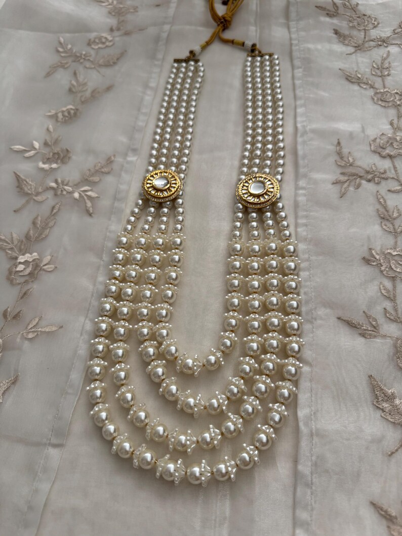 May include: A multi-strand pearl necklace with gold-toned circular accents. The necklace features multiple strands of white pearls, connected by decorative gold-colored clasps. The necklace is displayed on a white, textured surface.