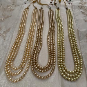 May include: Four strands of faux pearl necklaces in varying shades of cream and gold. Each necklace has three strands of pearls and is tied with a gold cord. The necklaces are displayed on a white, textured surface.