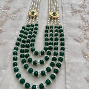 May include: A multi-strand necklace featuring alternating strands of white pearls and dark green beads. The necklace has gold-colored circular accents with a green center. The necklace is displayed on a white, textured surface.