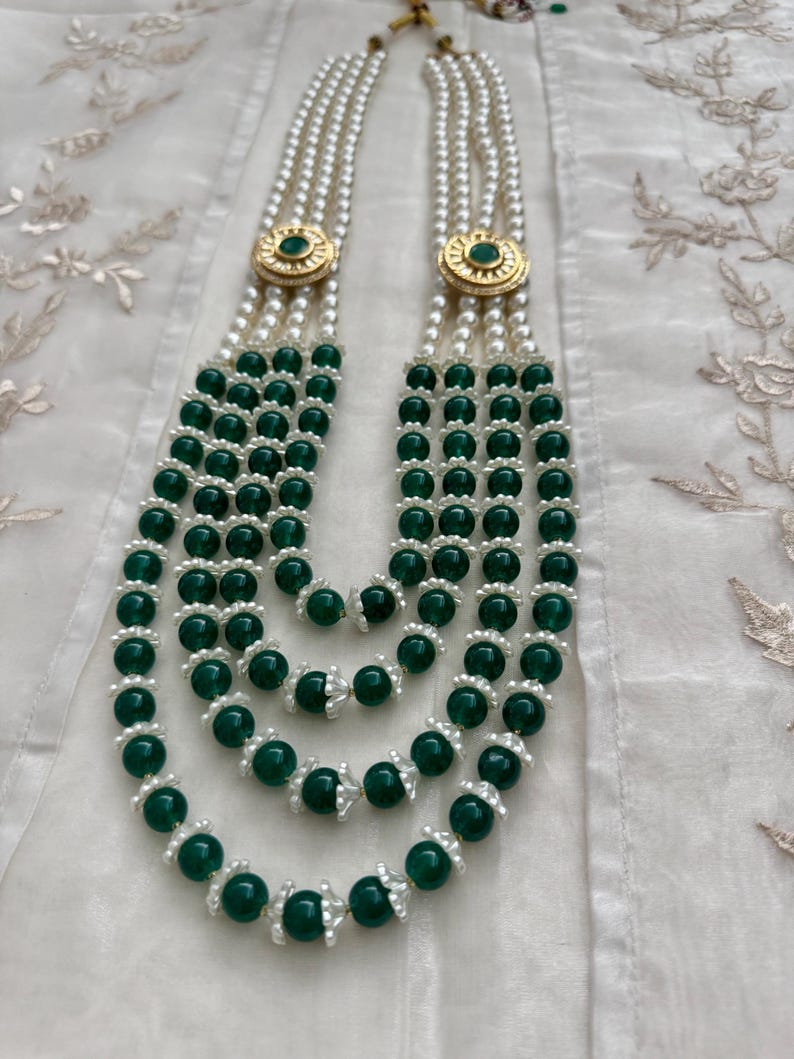 May include: A multi-strand necklace featuring alternating strands of white pearls and dark green beads. The necklace has gold-colored circular accents with a green center. The necklace is displayed on a white, textured surface.
