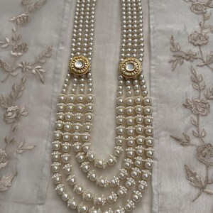 May include: A multi-strand pearl necklace with gold-toned circular accents. The necklace features multiple strands of white pearls, connected by two gold-colored circular clasps with a white center. The necklace is displayed on a white, textured surface.