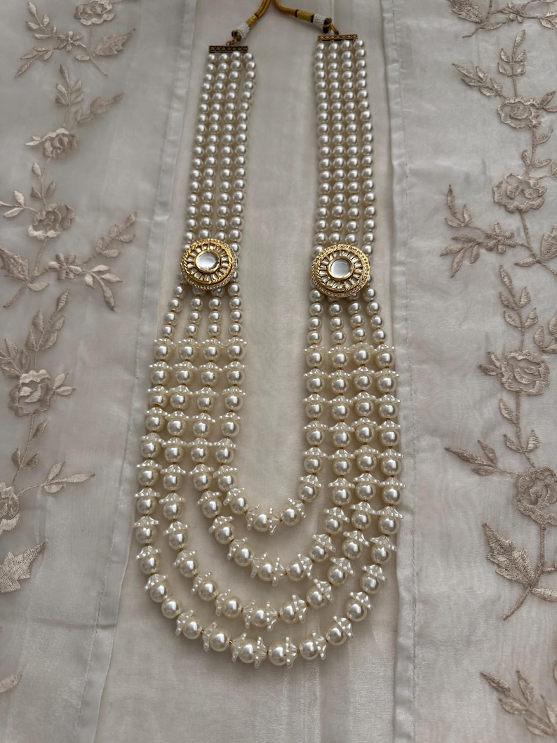 May include: A multi-strand pearl necklace with gold-toned circular accents. The necklace features multiple strands of white pearls, connected by two gold-colored circular clasps with a white center. The necklace is displayed on a white, textured surface.