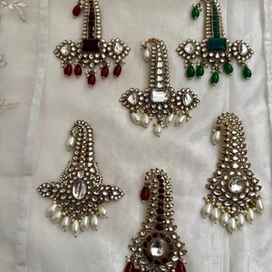 May include: A collection of gold-coloured earrings with detailed designs. Each earring has a central gemstone, surrounded by smaller stones and hanging teardrop beads in red, green, and white. Some have pearl embellishments. The earrings are displayed on a white surface.