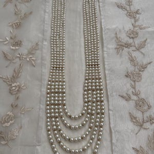 May include: A multi-strand pearl necklace with a gold clasp. The necklace features several strands of white pearls, graduating in length. The necklace is displayed on a white, textured surface with floral embroidery.