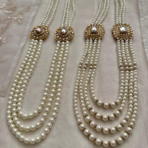 May include: Four strands of white pearl necklaces with gold-toned square and oval pendants. The pendants are encrusted with clear stones. The necklaces are displayed on a white, textured surface.