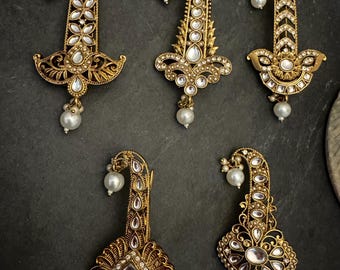 May include: Six ornate, gold-toned earrings with pearl accents are displayed against a dark background. Each earring features intricate filigree and is adorned with clear, sparkling stones. The earrings have a curved, elongated design.