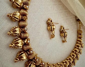 May include: A gold necklace and earrings set with a delicate design featuring small, ornate, teardrop-shaped pendants with clear stones. The necklace is made of round beads with a textured surface.