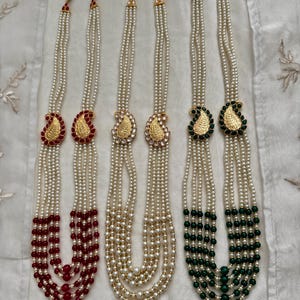 May include: Several pearl necklaces with gold pendants in a paisley design. The necklaces have multiple strands of white pearls, with some featuring red or green beads at the bottom. The pendants are gold with red or green accents.