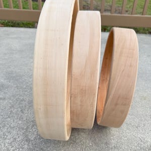 May include: Three unfinished wooden hoops of varying sizes, arranged in a vertical stack. The wood has a light, natural tone with visible grain. The hoops are likely for crafts or decorative purposes.