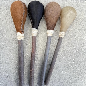 May include: Four handcrafted wooden clubs with different colored heads. The heads are covered in brown, black, and tan animal hide. The handles are made of wood and wrapped with a white material. The clubs are arranged vertically against a gray background.
